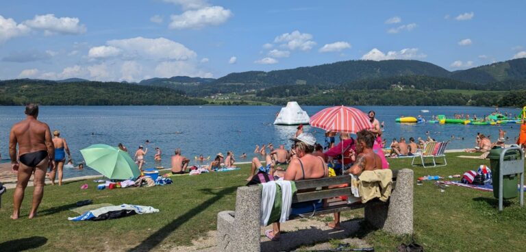 Blick auf den See Velenje mit Strand, Schwimmern und Bäumen